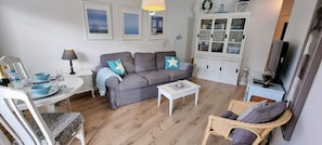A living room with a grey sofa, a white cabinet, a wicker chair, and a dining table with a blue bowl and glasses. - Ferienwohnung Nr. 33 (St. Peter-Ording)
