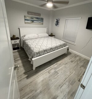 Un dormitorio con cama, mesita de noche, estante en la pared, ventana con persianas y ventilador de techo.