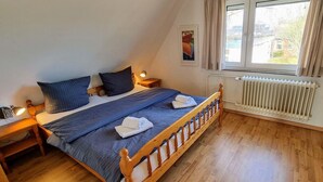 A bedroom with a wooden bed, a nightstand, a chair, a radiator, and a window with curtains. - Ferienhaus "deister" (St. Peter-Ording)