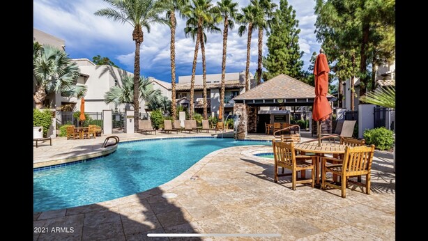 A swimming pool with a tiled deck, surrounded by palm trees and a covered seating area with wooden furniture. - Boutique Condo in North Scottsdale (Scottsdale)