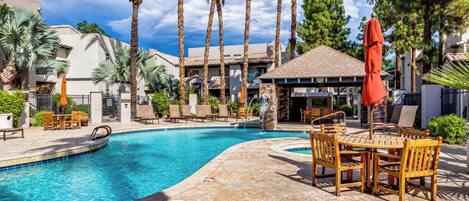 A swimming pool with a tiled deck, surrounded by palm trees and a covered seating area with wooden furniture.