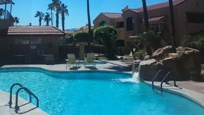 A swimming pool with clear blue water, surrounded by palm trees and a building with a tiled roof.