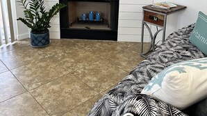 A living room with a flat-screen TV mounted above a white mantelpiece, a fireplace with three blue candles, a potted plant, and a patterned sofa with a teal pillow.