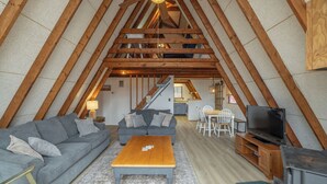 Een gezellige zolderkamer met een bank, salontafel en televisie, met zichtbare houten balken en een trap naar een keuken.