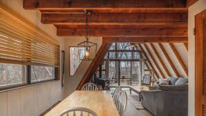 Een gezellig slaapkamer op zolder met houten plafond, een eettafel met stoelen, een grijze bank en een groot raam.