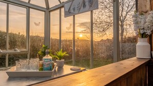 Eine Bar mit Blick auf den Sonnenuntergang durch große Fenster, einem Schild mit Pflanzenabbildungen und einer Tablett mit Gläsern und einer Flasche.