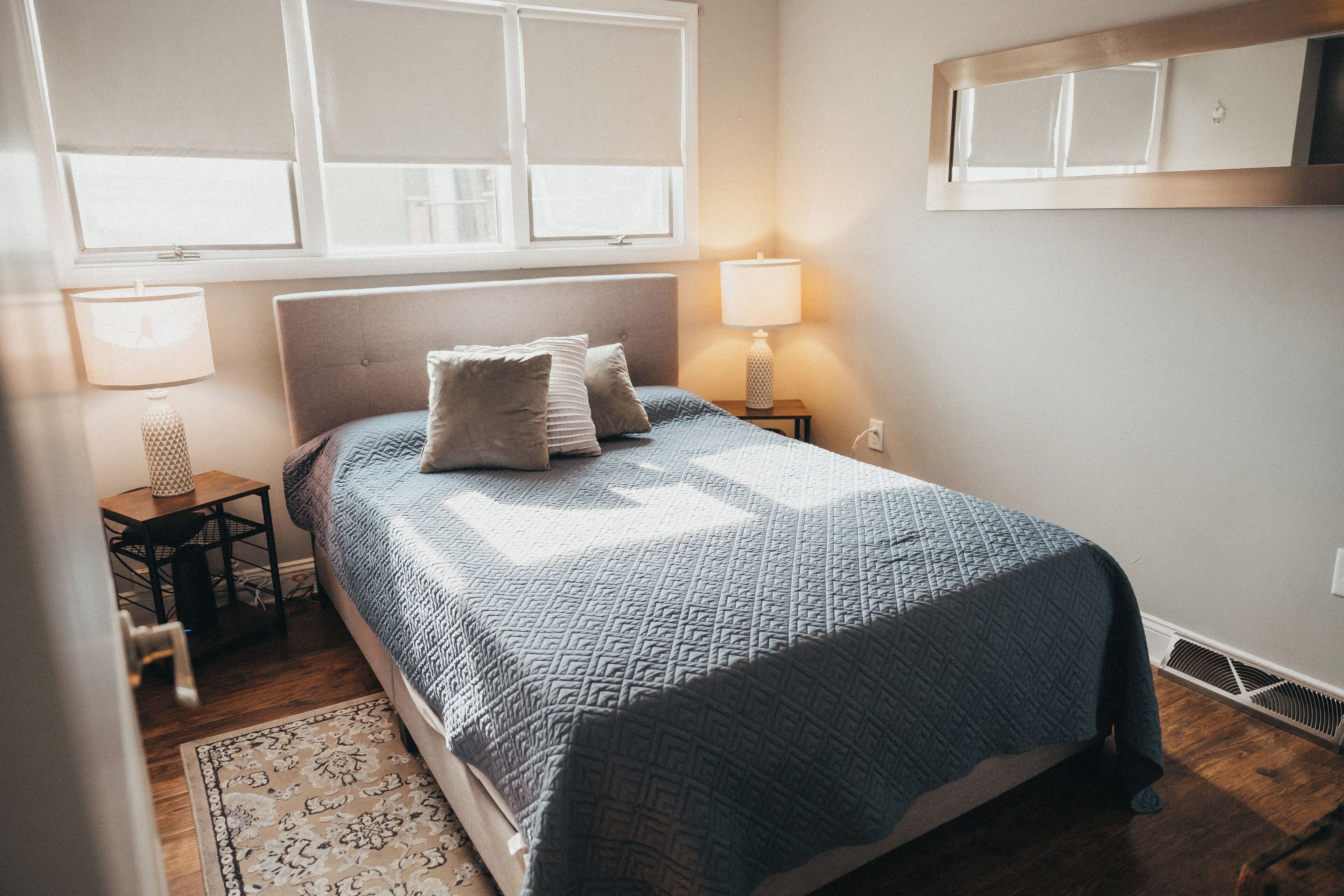 A bedroom with a bed, two bedside tables, a nightstand, a window, and a wall-mounted mirror.