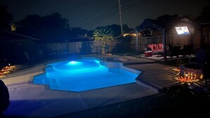 Une piscine nocturne avec un bord carrelé, entourée d’une clôture en bois, et un petit espace patio aménagé avec du mobilier d’extérieur.