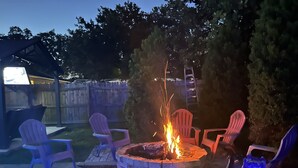 Un jardin avec un foyer entouré de fauteuils Adirondack, une clôture en bois et un téléviseur fixé sur une structure.