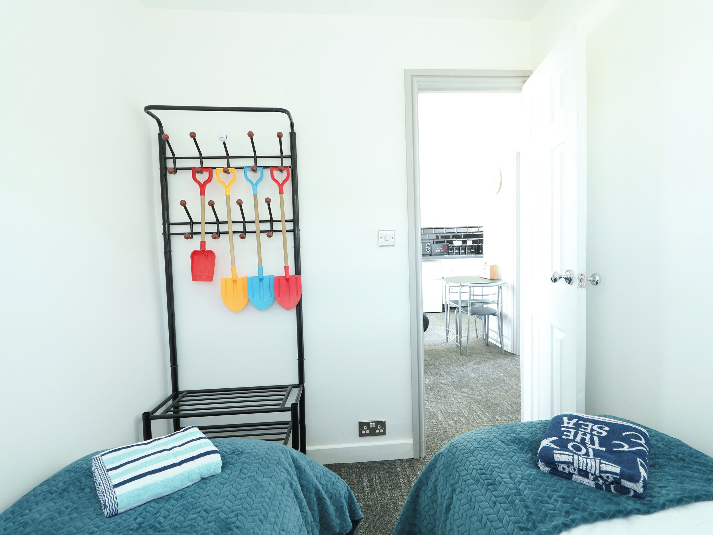 A bedroom with two beds, a metal rack with shovels, a kitchen in the background, and a door leading to another room.