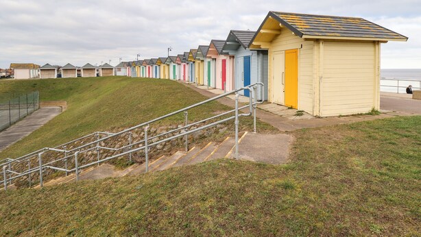 Une rangée de cabanes de plage aux couleurs vives, avec une colline verdoyante au premier plan et une étendue d’eau en arrière-plan.