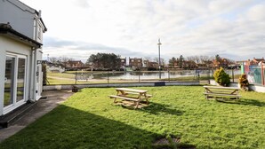 Un espace extérieur verdoyant, avec des tables de pique-nique, d’où l’on peut admirer un plan d’eau et des bâtiments en arrière-plan.