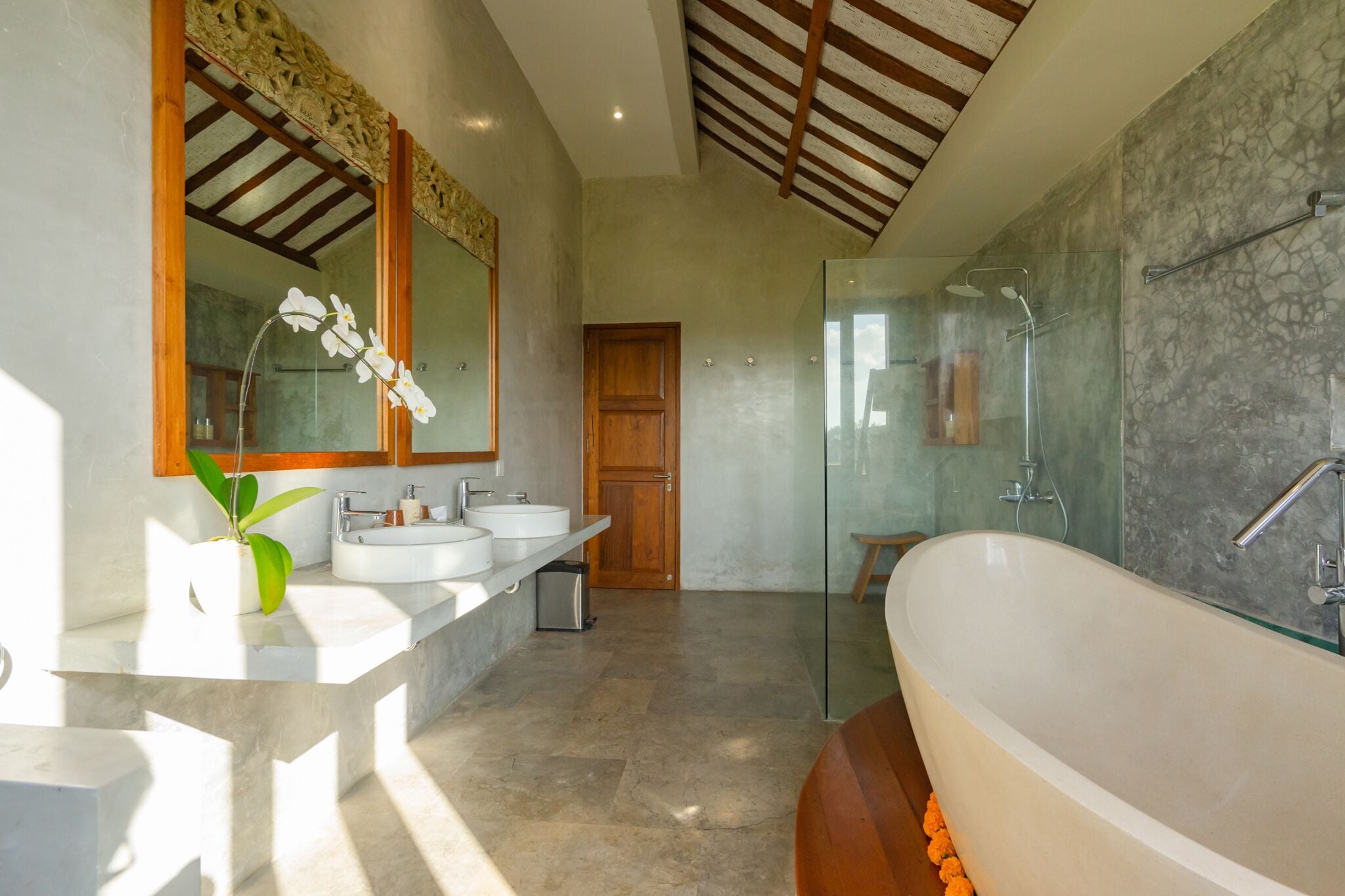 Un baño moderno con una bañera independiente grande, dos lavamanos, una ducha con cerramiento de vidrio y una puerta de madera.