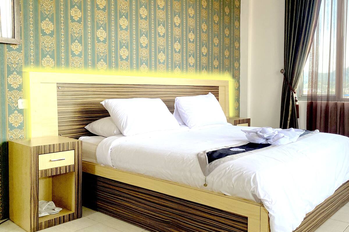 A neatly made bed with white linens and pillows, a wooden headboard, a bedside table with a drawer, and a window with curtains.