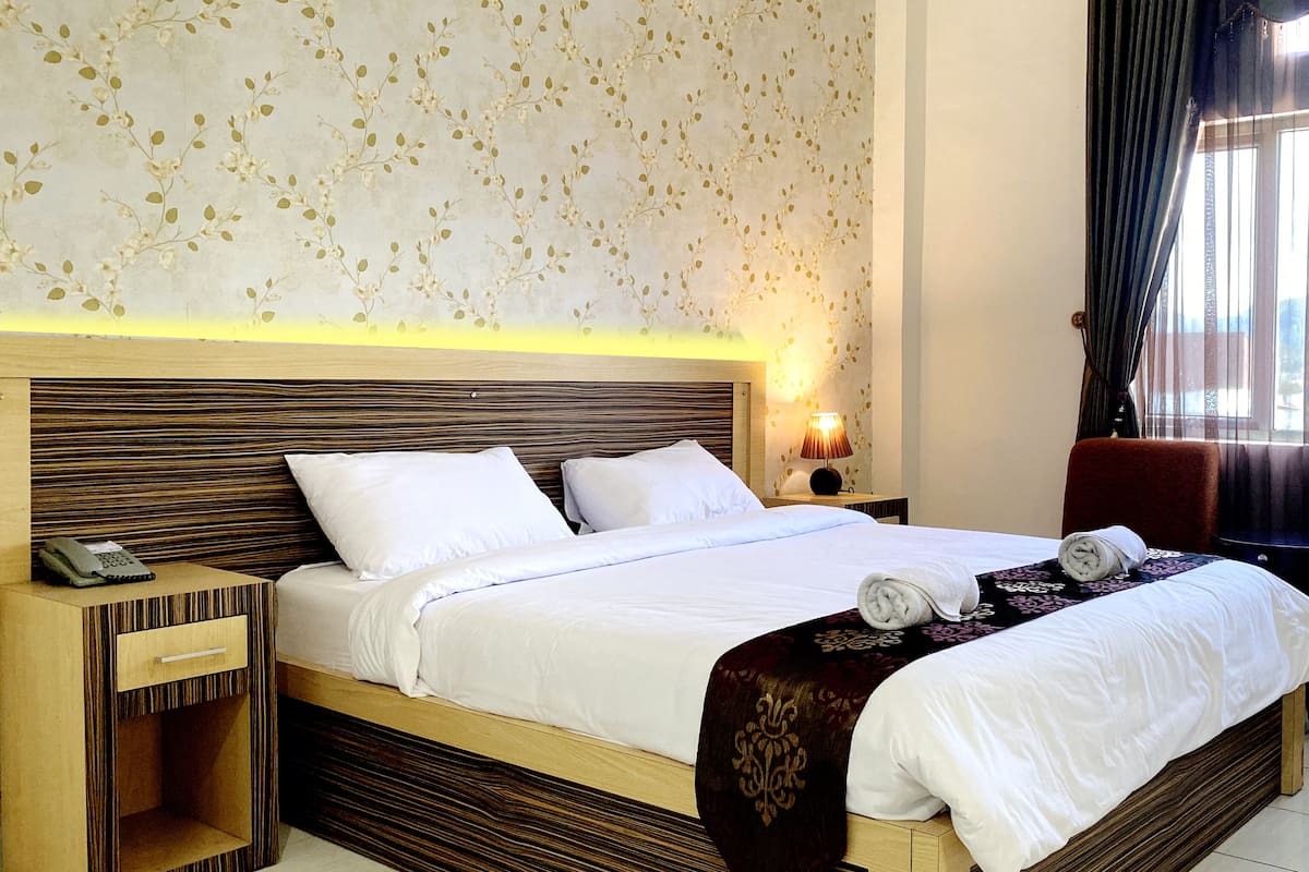 A hotel room with a large bed, a bedside table with a lamp, a chair, and a view of the outdoors through the window.