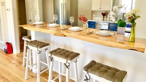 Una cocina moderna con encimera de mármol, armarios blancos y un refrigerador de acero inoxidable. Hay tres taburetes de bar con asientos acolchados y un florero con flores sobre la encimera.