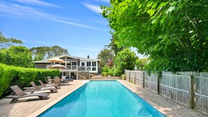 A swimming pool with lounge chairs and umbrellas, surrounded by a wooden fence and lush greenery. -  Southampton Gem House with Beautiful Heated Pool (Southampton)