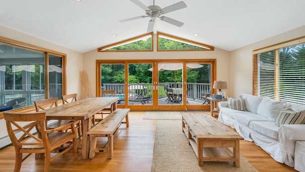 A spacious living room with a wooden dining table, benches, a white sofa, and a ceiling fan. -  Southampton Gem House with Beautiful Heated Pool (Southampton)