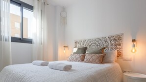 Un lit bien fait, avec des serviettes pliées, une tête de lit décorée, deux appliques murales et une petite table de chevet.