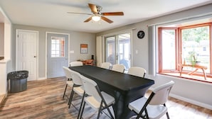 A dining area with a long table, white chairs, a ceiling fan, and large windows with a view of the outside.