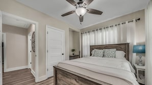 Un dormitorio con una cama de madera, un ventilador de techo, una mesita de noche con una lámpara y una puerta que da a otra habitación.