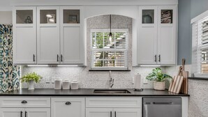 Cocina con armarios blancos, pileta de acero inoxidable y ventana con persianas.