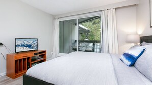 A bedroom with a bed, a TV on a wooden stand, a balcony with a chair, and a view of trees.