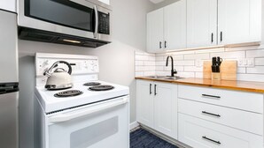 A modern kitchen with white cabinets, a stainless steel microwave, a kettle on the stove, and a wooden countertop.