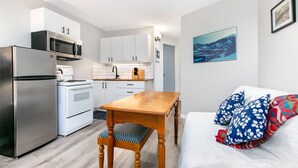 A compact kitchen with white cabinets, a microwave, a stove, and a refrigerator. There's a wooden table with a patterned cushion, a white sofa with blue and red pillows, and a framed picture on the wall.