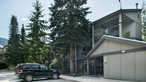A modern building with a garage, a black SUV parked in front, and tall evergreen trees.