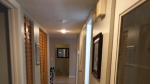 Um corredor estreito, com piso de azulejos, possui uma porta à direita, um espelho na parede e um quadro emoldurado acima da porta.