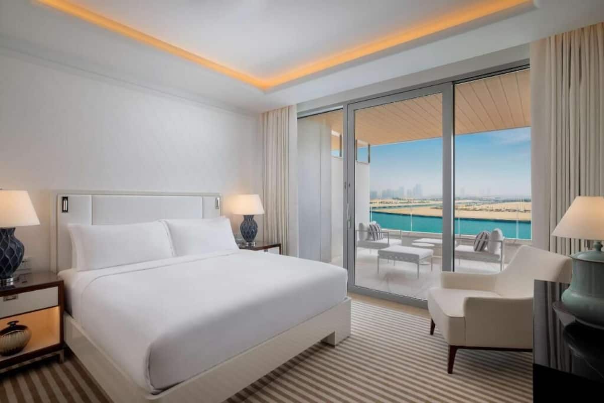A hotel room with a large bed, two bedside tables, a chair, a balcony with a view of the beach and city skyline.