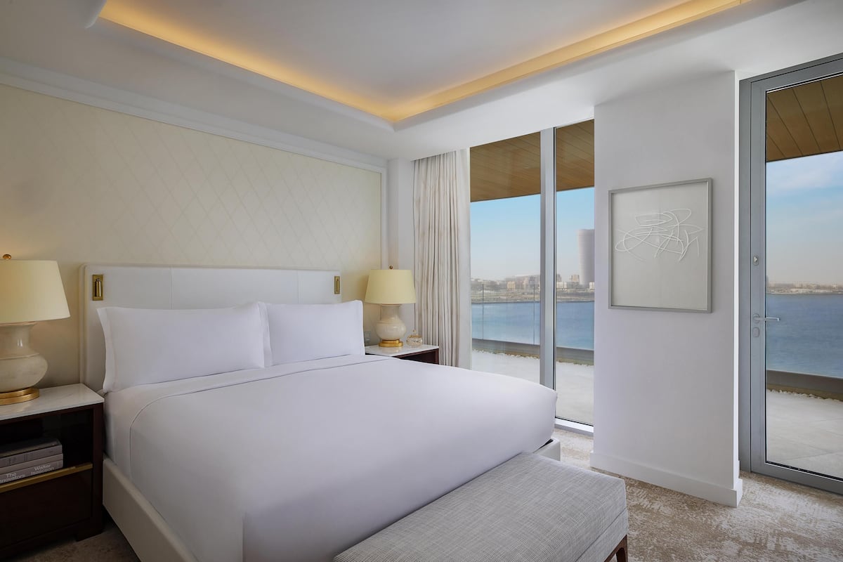 A hotel room with a large bed, two bedside lamps, a view of the sea, and a sliding glass door.