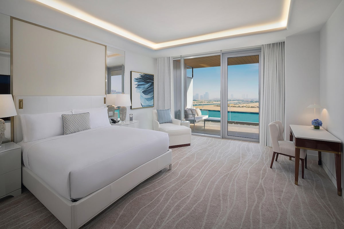 A modern hotel room with a large bed, a desk, a chair, a balcony with a view of the beach, and a cityscape.