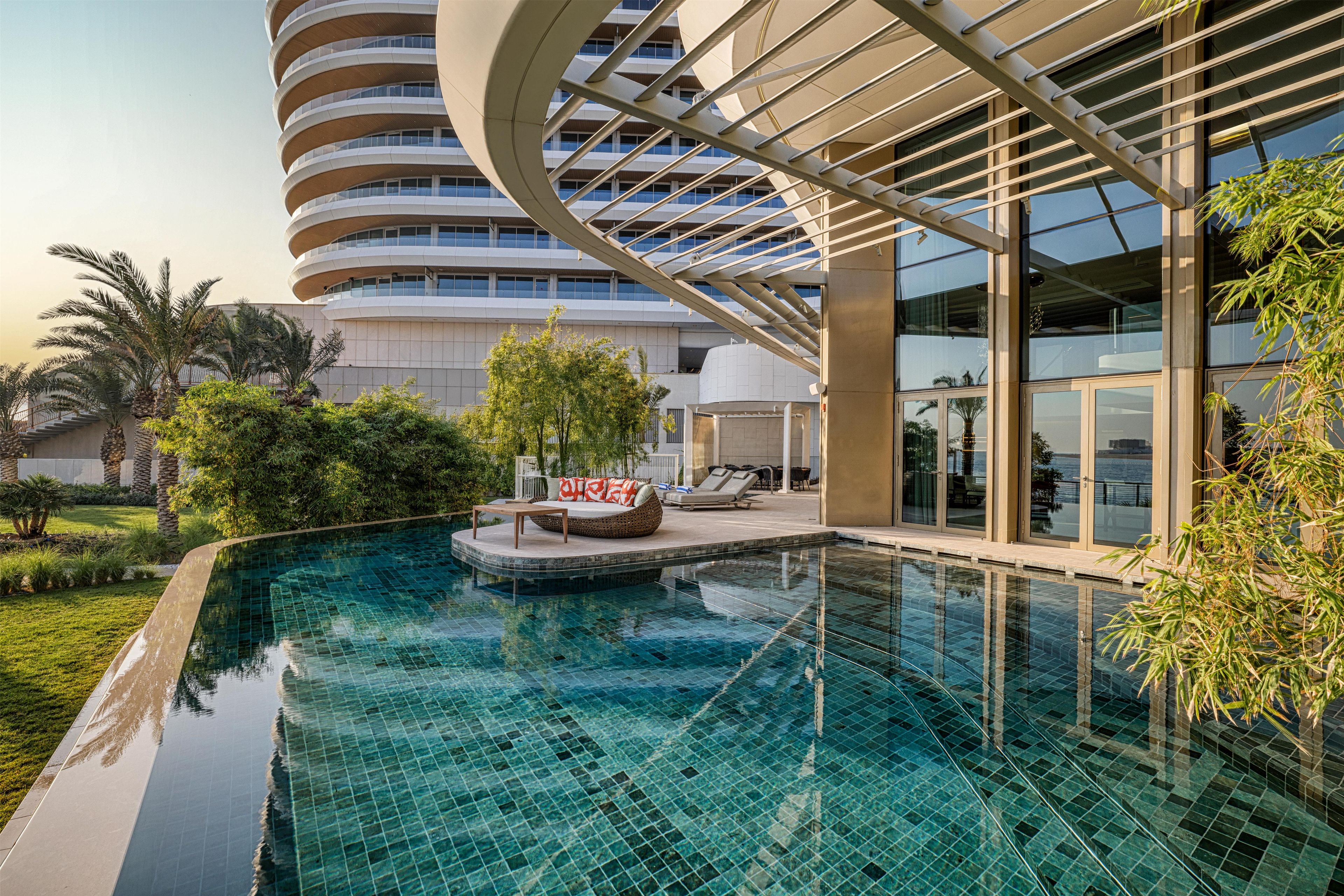 Un bâtiment moderne à la façade incurvée, doté d’une piscine avec un espace salon aménagé en contrebas et entouré d’une végétation luxuriante.