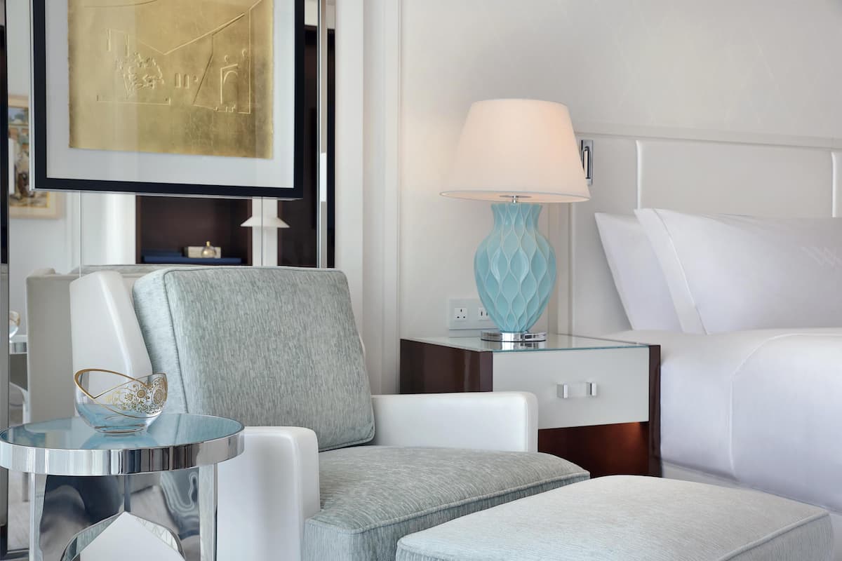 A hotel room with a bed, a chair, a small table with a lamp, and a framed picture on the wall.