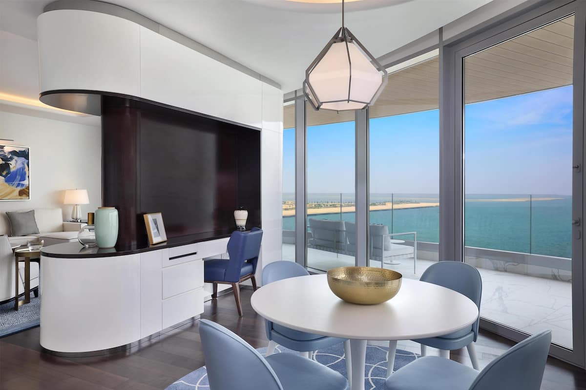 A modern living room with a curved white console, a flat-screen TV, a dining area with a round table and blue chairs, and a balcony with a view of the sea.