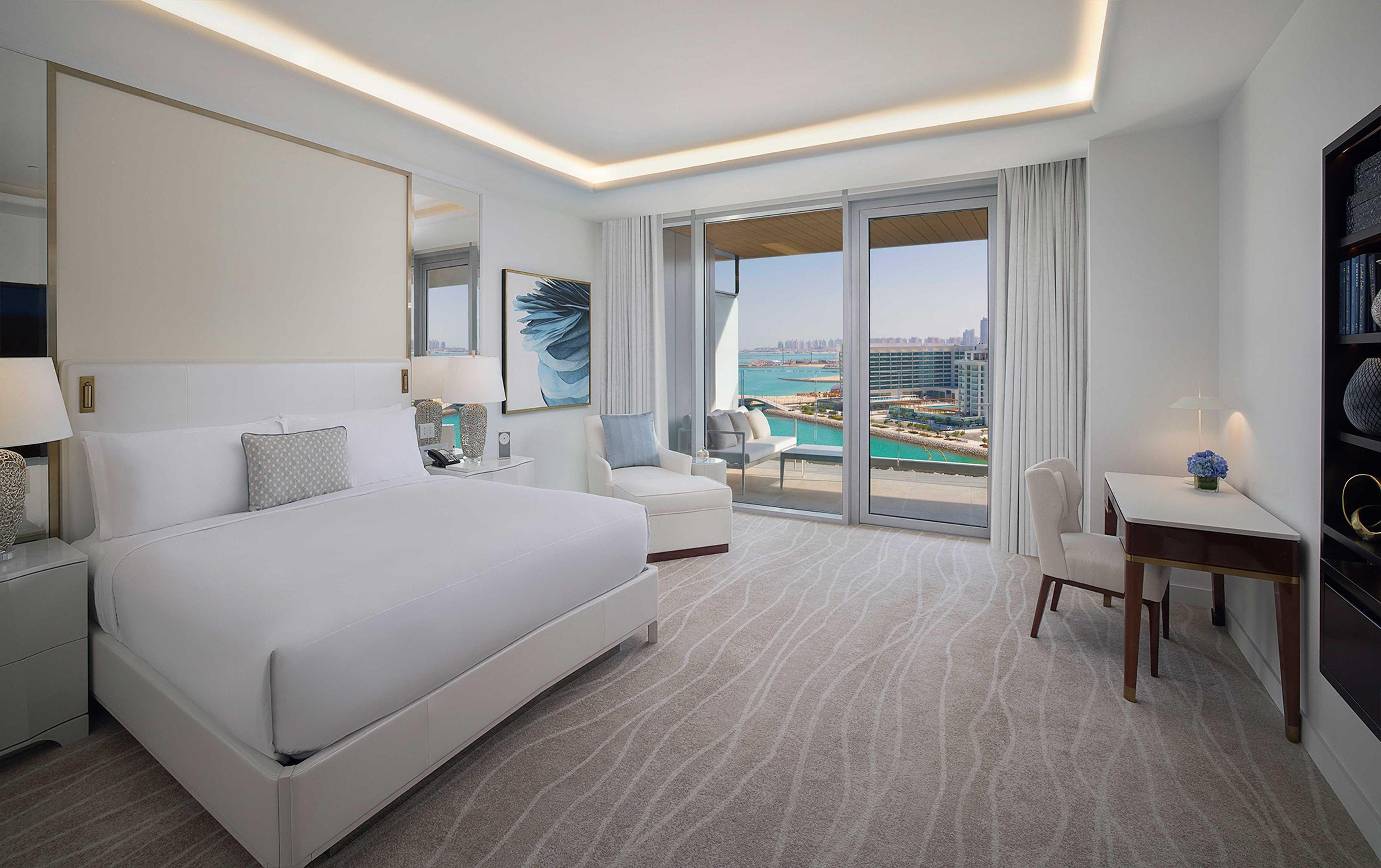 Habitación de hotel con una cama grande, un escritorio, una silla, una mesita, una lámpara, una estantería y un ventanal con vistas a la ciudad y al agua.