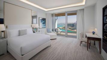 Habitación de hotel con una cama grande, un escritorio, una silla, una mesita, una lámpara, una estantería y un ventanal con vistas a la ciudad y al agua.