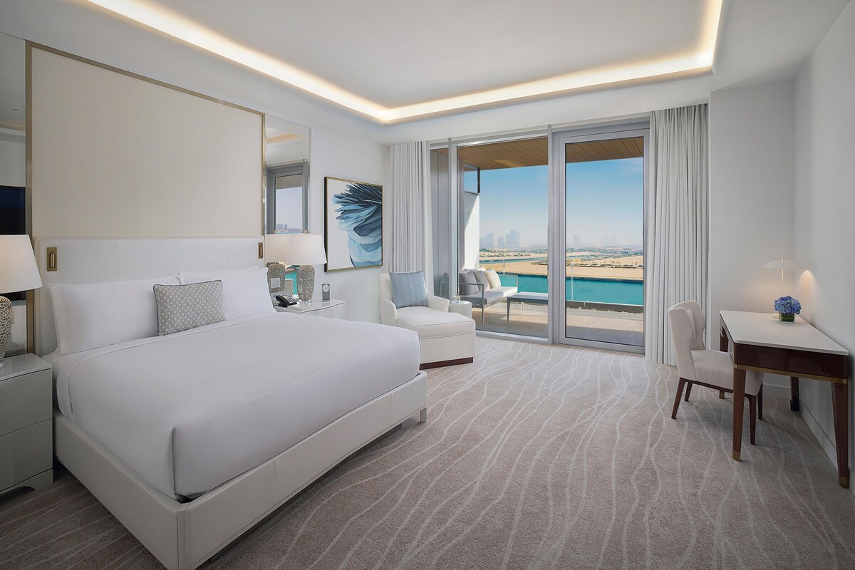 A modern hotel room with a large bed, a desk, a chair, a balcony with a view of the beach, and a cityscape.