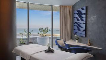 Enjoy a panoramic sea view from the large window in this stylish room, featuring a bed, massage table, and cozy seating area.