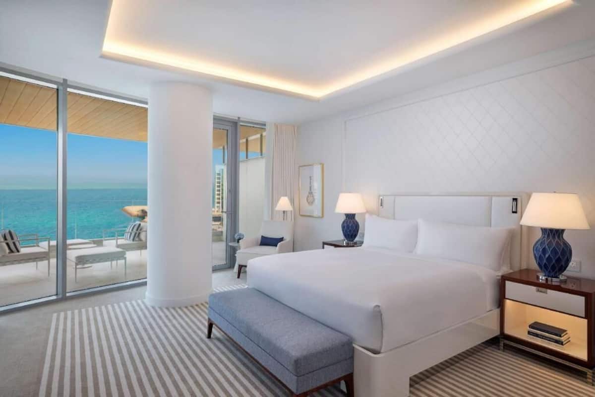 A modern hotel room with a large bed, bedside tables, a bench, and a view of the ocean.