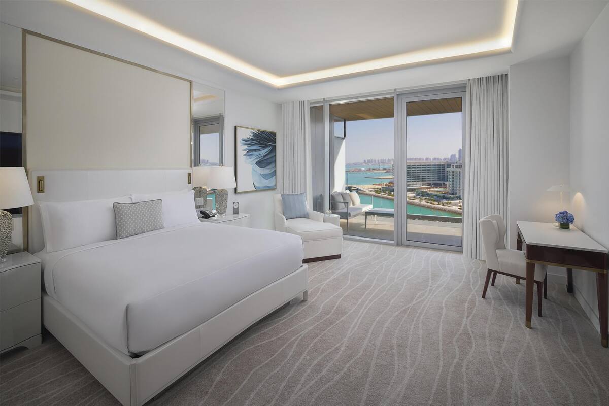 A hotel room with a large bed, a desk, a chair, a sofa, a large window with a city view, and a balcony.