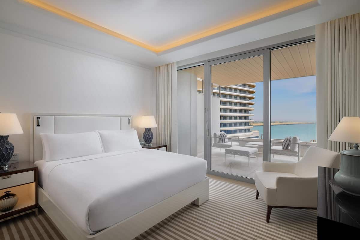 A hotel room with a large bed, two bedside tables with lamps, a chair, a balcony with a view of the sea, and a glass door leading to a pool area.