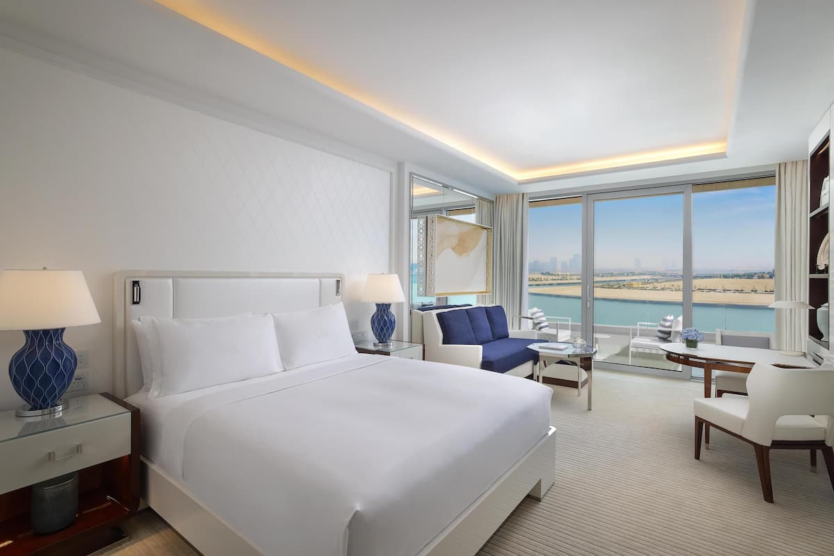A hotel room with a large bed, a seating area with a view of the beach, and a balcony with a cityscape view.