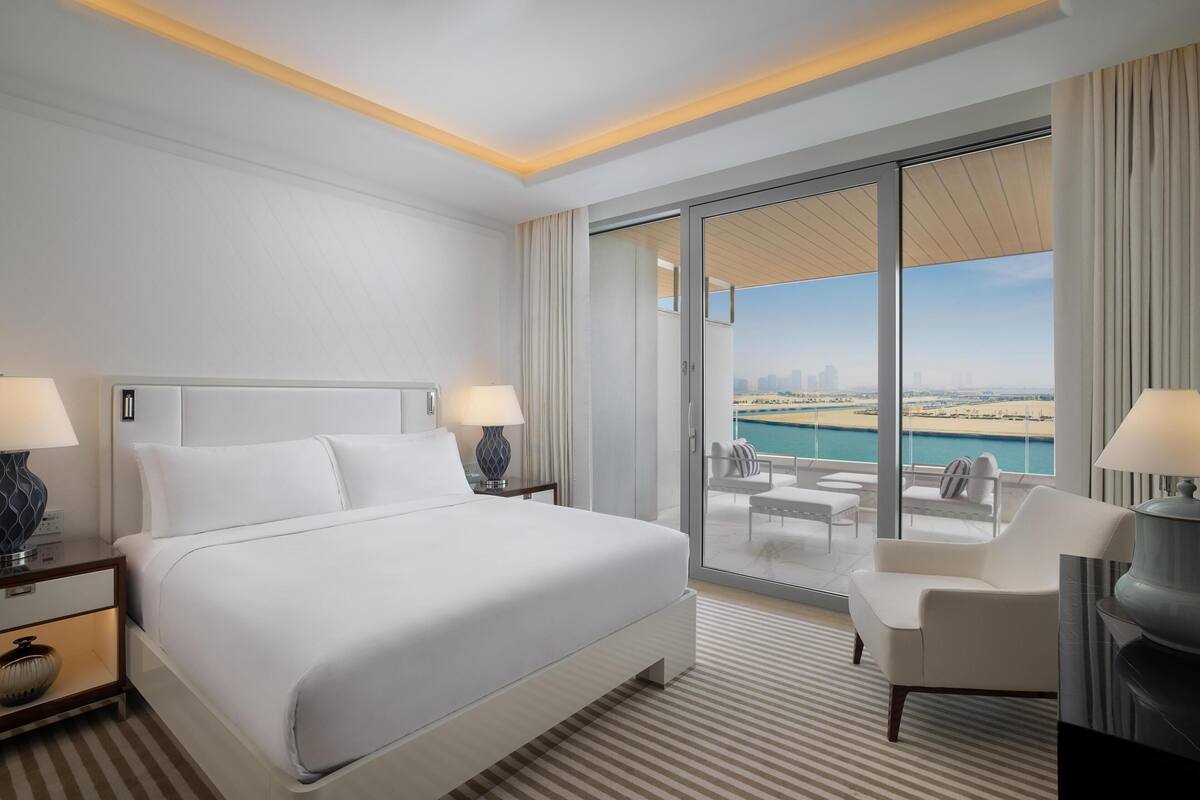 A modern hotel room with a large bed, two bedside tables, a chair, a balcony with a view of the beach and city skyline.