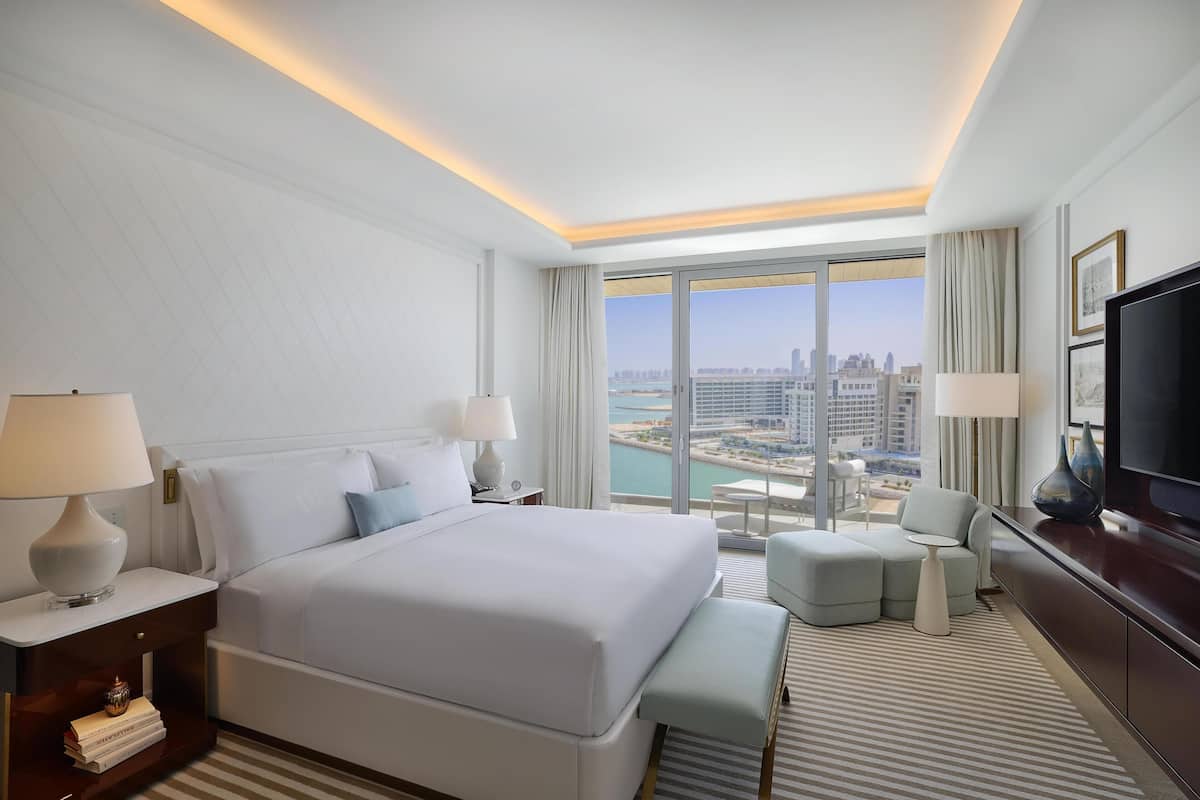 A hotel room with a large bed, a TV, a desk with a lamp, and a view of a cityscape.