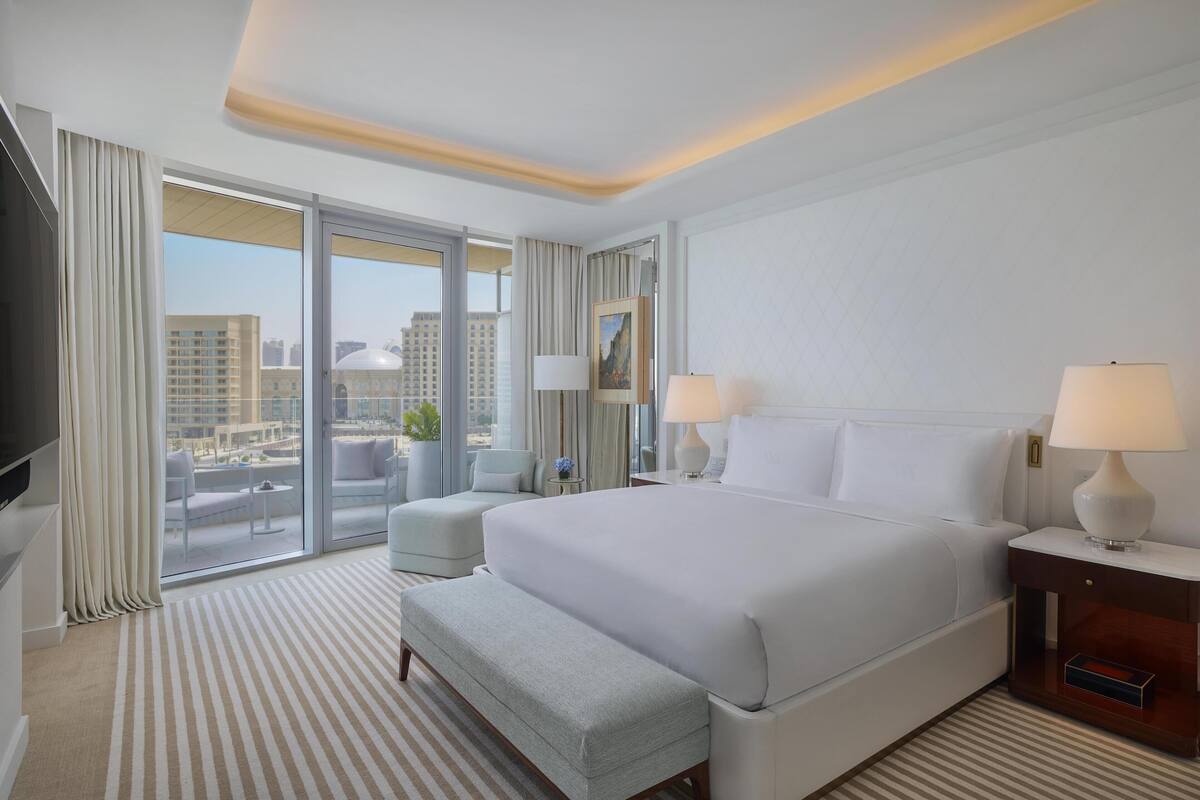 A hotel room with a large bed, two bedside tables with lamps, a flat-screen TV, a sofa, and a large window with a city view.