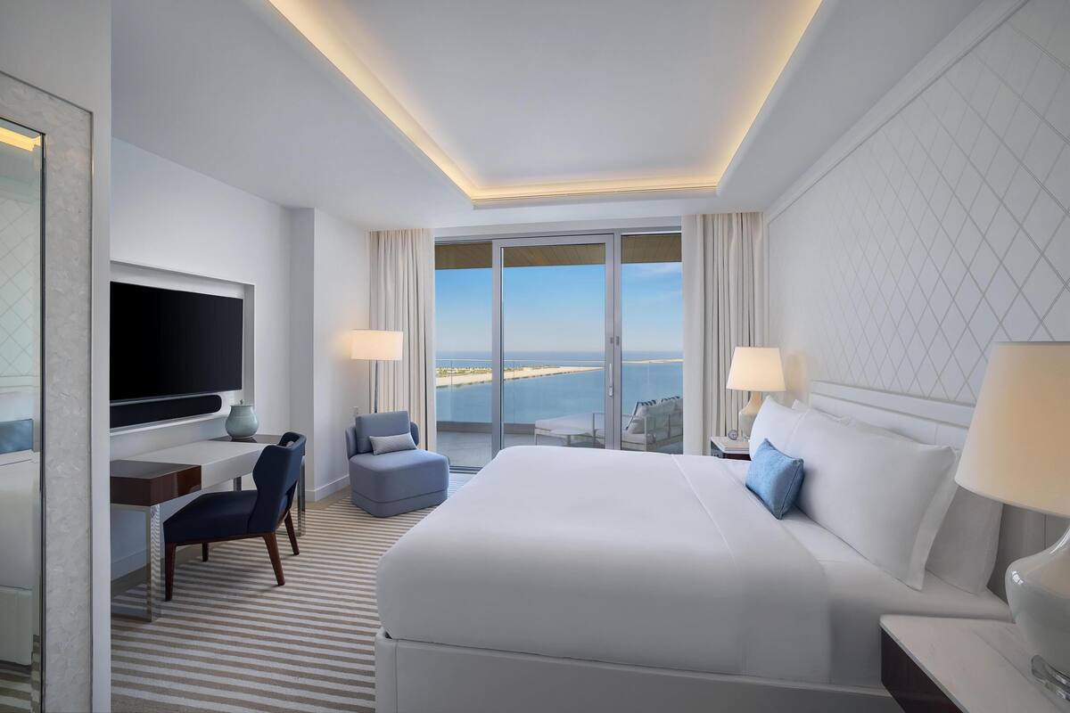 A hotel room with a large bed, a desk, a chair, a TV, and a balcony with a view.