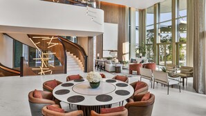 A spacious lobby with a central round table, seating arrangements, a staircase, and large windows.